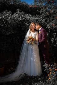 A couple in wedding attire poses affectionately in a garden setting. The bride wears a white gown with a long veil and holds a bouquet of sunflowers, while the groom dons a dark suit with a boutonniere. They are surrounded by lush greenery, and there are small orange flowers in the foreground.