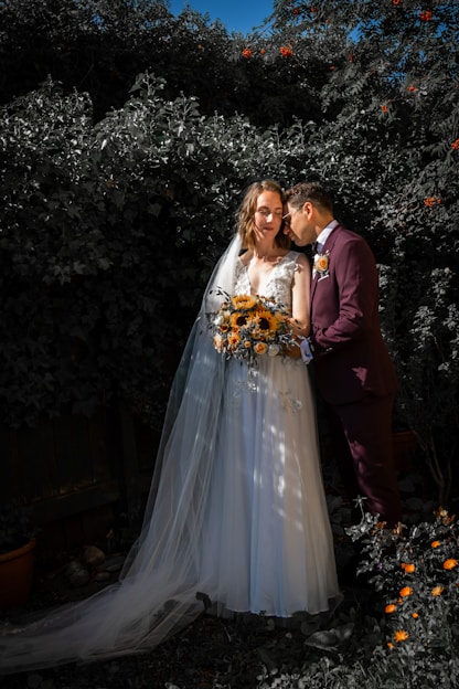 A couple in wedding attire poses affectionately in a garden setting. The bride wears a white gown with a long veil and holds a bouquet of sunflowers, while the groom dons a dark suit with a boutonniere. They are surrounded by lush greenery, and there are small orange flowers in the foreground.