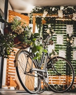 A vintage bicycle is adorned with a bouquet of lush greenery and white flowers, positioned in a bright, warmly lit indoor setting. The background features decorative green foliage and strings of small lights, partially obscuring wooden panels and sheets of hanging paper.