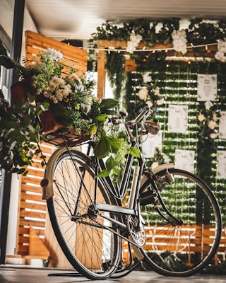 A vintage bicycle is adorned with a bouquet of lush greenery and white flowers, positioned in a bright, warmly lit indoor setting. The background features decorative green foliage and strings of small lights, partially obscuring wooden panels and sheets of hanging paper.