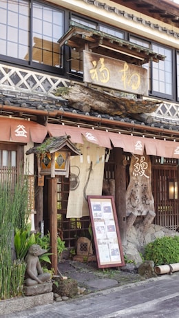 A traditional Japanese storefront features wooden architecture with detailed carvings and a large hanging wooden sign with Japanese characters. The entrance is adorned with potted plants, stone decorations, and a small statue. Fabric banners hang beside a framed menu or information board, all contributing to an inviting and rustic aesthetic.
