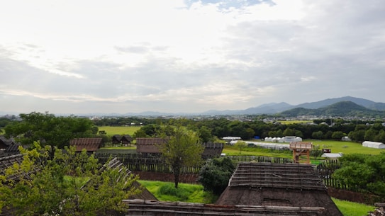 An expansive landscape featuring a series of thatched-roof structures in the foreground, surrounded by lush greenery and trees. In the distance, low-lying mountains rise against a partly cloudy sky. The horizon separates the vast green fields from the clear blue sky.