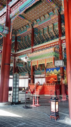 Richly decorated interior of a traditional Korean palace with intricate colorful patterns on the wooden beams and ceiling. The room features carved wooden columns, ornate painting, and traditional lamps. Dominating the room is an elevated red throne set against a backdrop of a vibrant blue mural with mountain motifs.