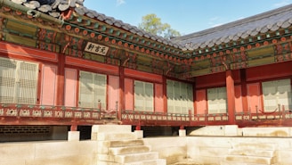A traditional Asian architectural structure featuring ornate wooden detailing and intricate patterns. The building has a sloping tiled roof and shutters, situated on a stone platform. Rich, earthy colors dominate the structure, with decorative elements adding cultural significance.