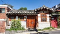 A traditional Korean hanok house with a curved tiled roof and wooden door. The building features wooden lattice windows and a stone wall at the base. There is greenery, including bamboo, in front of the house, and utility wires are visible above.