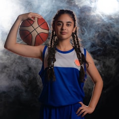 A young basketball player with braided hair is standing confidently, holding a basketball against her head with her left hand. She is wearing a blue and white basketball uniform. The background is smoky with bright light coming from behind.