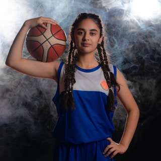 A young basketball player with braided hair is standing confidently, holding a basketball against her head with her left hand. She is wearing a blue and white basketball uniform. The background is smoky with bright light coming from behind.