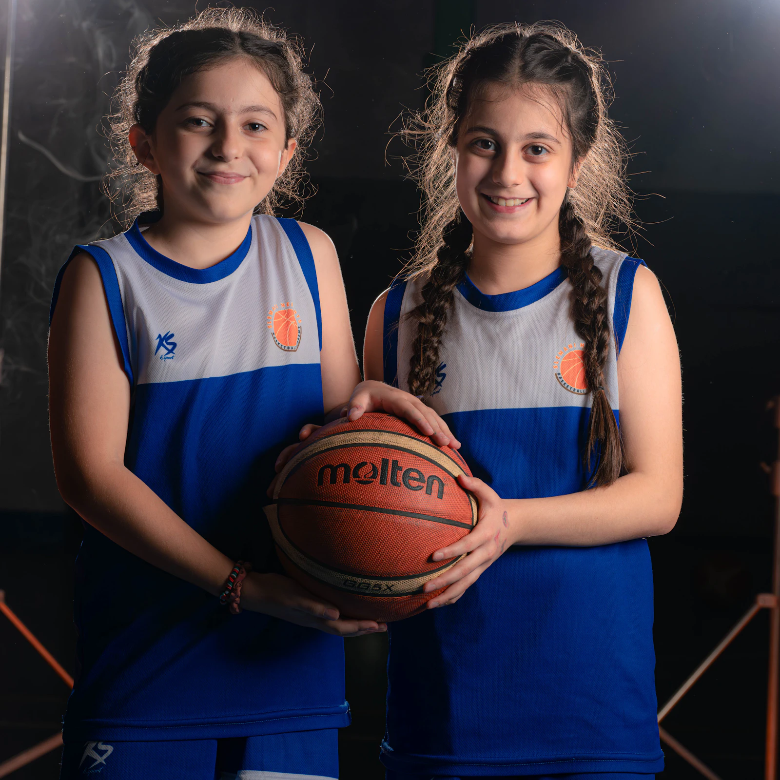 Two teen girls in matching blue and white basketball uniforms holding a basketball together on the court.
