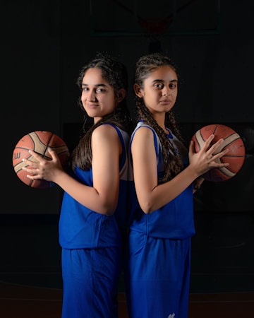 Two young basketball players stand back-to-back in blue uniforms, each holding a basketball under one arm. They both have braided hair and are posed confidently against a dark background, with an unfocused basketball hoop visible in the top background area.