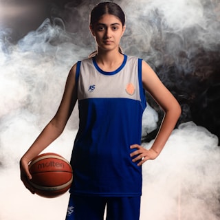 A person wearing a blue and white basketball jersey stands confidently, holding a basketball in one hand. Smoke or mist surrounds the background, adding a dramatic effect.