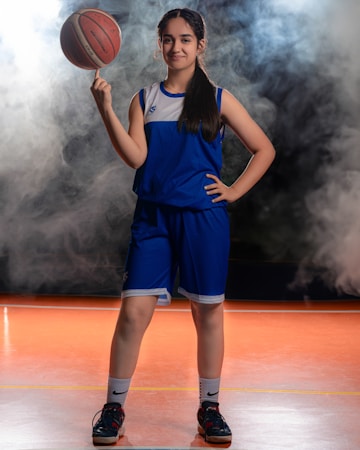 A young person is confidently posing on a basketball court, wearing a blue sports uniform and holding a basketball poised on one finger. The background is filled with a misty or smoky atmosphere, creating a dynamic and energetic setting.