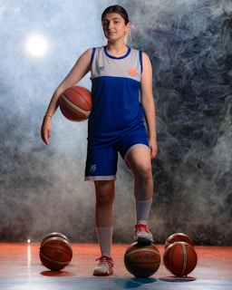 A person is standing with a confident pose, dressed in a blue and white sports uniform. They are holding a basketball under their right arm while their left foot is resting on another basketball. There are three additional basketballs placed around on the ground. The background is smoky, giving a dramatic effect, with light shining through the smoke.