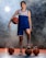 A person is standing with a confident pose, dressed in a blue and white sports uniform. They are holding a basketball under their right arm while their left foot is resting on another basketball. There are three additional basketballs placed around on the ground. The background is smoky, giving a dramatic effect, with light shining through the smoke.
