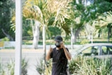 A person, partially obscured by reflections, is taking a photograph through a glass window. They are wearing a dark shirt and cap and giving a thumbs-up gesture. In the background, there are blurred views of palm trees, a parked car, and a road.