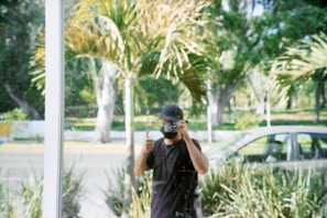 A person, partially obscured by reflections, is taking a photograph through a glass window. They are wearing a dark shirt and cap and giving a thumbs-up gesture. In the background, there are blurred views of palm trees, a parked car, and a road.