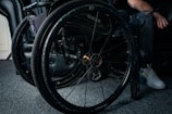 A close-up view of a wheelchair showing detailed parts of its wheels and frame. The wheelchair is positioned on a carpeted floor next to a black leather chair. A person's hand rests on the armrest while one leg is visible, wearing jeans and white sneakers.