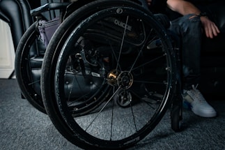 A close-up view of a wheelchair showing detailed parts of its wheels and frame. The wheelchair is positioned on a carpeted floor next to a black leather chair. A person's hand rests on the armrest while one leg is visible, wearing jeans and white sneakers.