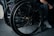 A close-up view of a wheelchair showing detailed parts of its wheels and frame. The wheelchair is positioned on a carpeted floor next to a black leather chair. A person's hand rests on the armrest while one leg is visible, wearing jeans and white sneakers.