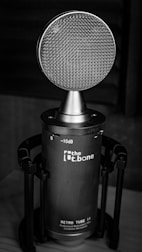 A close-up of a microphone with a retro design placed on a stand. The microphone features a large round mesh grille and a cylindrical body with branding and decibel markings visible on the surface.