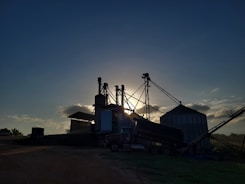 Silhouetted structures of a grain processing facility are seen against a setting or rising sun, creating a dramatic effect. The sky is partially covered with soft clouds illuminated by the sunlight, and the ground surrounding the structures is mostly in shadow.