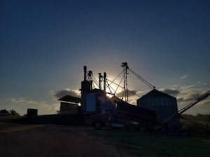 Silhouetted structures of a grain processing facility are seen against a setting or rising sun, creating a dramatic effect. The sky is partially covered with soft clouds illuminated by the sunlight, and the ground surrounding the structures is mostly in shadow.