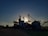 Silhouetted structures of a grain processing facility are seen against a setting or rising sun, creating a dramatic effect. The sky is partially covered with soft clouds illuminated by the sunlight, and the ground surrounding the structures is mostly in shadow.