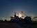 Silhouetted structures of a grain processing facility are seen against a setting or rising sun, creating a dramatic effect. The sky is partially covered with soft clouds illuminated by the sunlight, and the ground surrounding the structures is mostly in shadow.