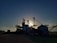 Silhouetted structures of a grain processing facility are seen against a setting or rising sun, creating a dramatic effect. The sky is partially covered with soft clouds illuminated by the sunlight, and the ground surrounding the structures is mostly in shadow.