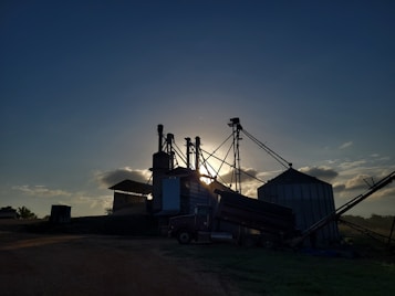 Silhouetted structures of a grain processing facility are seen against a setting or rising sun, creating a dramatic effect. The sky is partially covered with soft clouds illuminated by the sunlight, and the ground surrounding the structures is mostly in shadow.