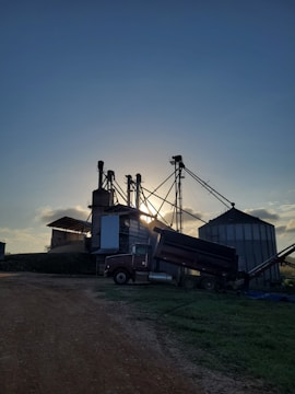 A rural industrial setting featuring agricultural storage structures, likely grain silos, with tall, cylindrical silos and conveyer systems against a backdrop of a setting or rising sun. A large truck is parked near the silos, with some equipment attached to it. The sky is clear with a few clouds, and the lighting suggests early morning or late afternoon.
