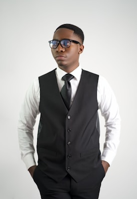 A person wearing a white dress shirt, black tie, and a black vest, standing with hands in pockets. They have short hair and are wearing glasses, with a neutral expression on their face.