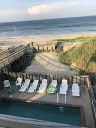 A scenic view overlooking a sandy beach and ocean, bordered by a wooden fence with a pathway leading to the beach. Multiple white lounge chairs are arranged around a small private pool, and a green table with a white chair is nearby. The area is surrounded by sand dunes and grass.