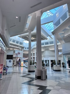 A spacious, modern shopping mall interior featuring tall columns, a high ceiling with skylights, and various shops lining the walkway. The area is well-lit with natural and artificial light, and there are planters with greenery positioned between columns. Several people can be seen walking in the distance.