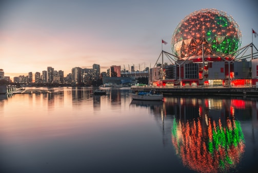 A stunning geodesic dome structure is illuminated with red and green lights and reflected on the calm waters of a nearby body of water. The backdrop features a city skyline as the sun sets, casting a warm glow across the sky.