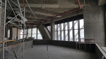 A partially constructed building interior with exposed concrete walls and large windows. Scaffolding and construction materials are present, indicating ongoing construction work. The ceiling shows exposed metal beams and pipes.