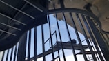 An architectural view featuring metal beams and railings with a clear sky visible through the gaps. The structural elements form a pattern of vertical and curved lines that create a sense of depth and perspective.