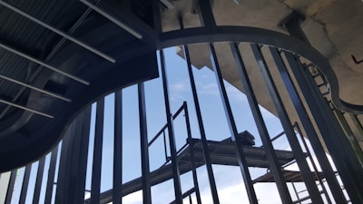 An architectural view featuring metal beams and railings with a clear sky visible through the gaps. The structural elements form a pattern of vertical and curved lines that create a sense of depth and perspective.