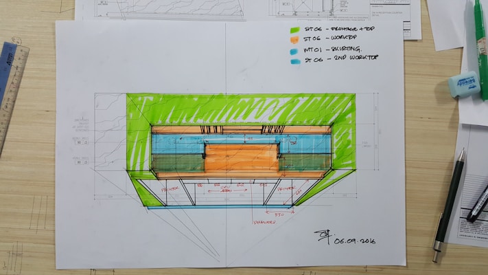 An architectural sketch appears on a desk, featuring a colored floorplan with measurements. Various tools and drawing supplies, including pens, a ruler, and an eraser, surround the plan. The drawing includes notes and uses vivid colors to highlight different sections.