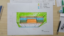 An architectural sketch appears on a desk, featuring a colored floorplan with measurements. Various tools and drawing supplies, including pens, a ruler, and an eraser, surround the plan. The drawing includes notes and uses vivid colors to highlight different sections.