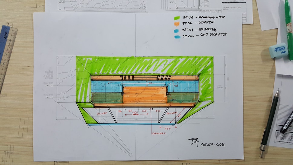 An architectural sketch appears on a desk, featuring a colored floorplan with measurements. Various tools and drawing supplies, including pens, a ruler, and an eraser, surround the plan. The drawing includes notes and uses vivid colors to highlight different sections.