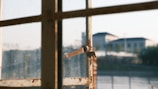 A sunlit, slightly rusty window latch on a multi-pane window with an out-of-focus view of buildings in the background.