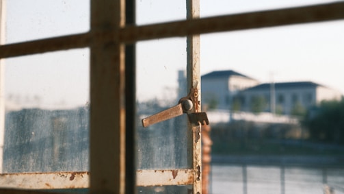 A sunlit, slightly rusty window latch on a multi-pane window with an out-of-focus view of buildings in the background.