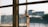 A sunlit, slightly rusty window latch on a multi-pane window with an out-of-focus view of buildings in the background.