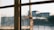 A sunlit, slightly rusty window latch on a multi-pane window with an out-of-focus view of buildings in the background.
