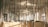 Tall bamboo stalks are arranged vertically in an indoor space, creating a forest-like atmosphere. Natural light from large windows illuminates the area, casting soft shadows on the walls and floor. Decorative panels with Asian calligraphy are positioned among the bamboo, adding an artistic and cultural element.