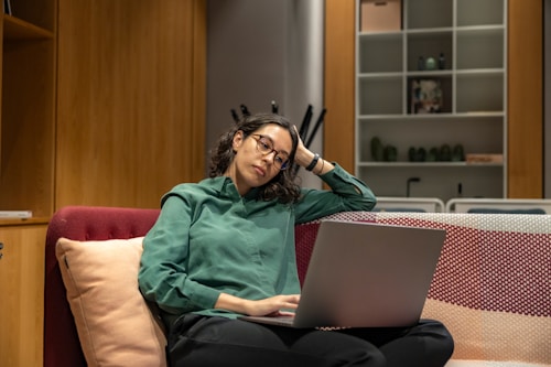 A person with glasses wearing a green shirt sits on a sofa with a laptop in their lap. They appear thoughtful or tired, with one hand resting on their head. The background includes wooden shelves with books and decorative items.