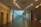 A modern, well-lit hallway with a polished dark tile floor and numerous ceiling lights. There are large stone walls on the left and a glass railing on the right leading to a metallic art installation. The receptionist desk on the right side has a flower arrangement, and the entire space feels spacious with its open design and light-colored walls.