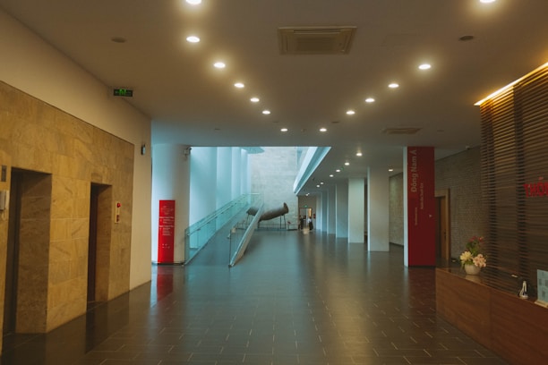 A modern, well-lit hallway with a polished dark tile floor and numerous ceiling lights. There are large stone walls on the left and a glass railing on the right leading to a metallic art installation. The receptionist desk on the right side has a flower arrangement, and the entire space feels spacious with its open design and light-colored walls.