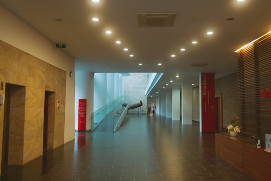 A modern, well-lit hallway with a polished dark tile floor and numerous ceiling lights. There are large stone walls on the left and a glass railing on the right leading to a metallic art installation. The receptionist desk on the right side has a flower arrangement, and the entire space feels spacious with its open design and light-colored walls.