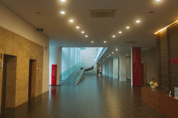 A modern, well-lit hallway with a polished dark tile floor and numerous ceiling lights. There are large stone walls on the left and a glass railing on the right leading to a metallic art installation. The receptionist desk on the right side has a flower arrangement, and the entire space feels spacious with its open design and light-colored walls.